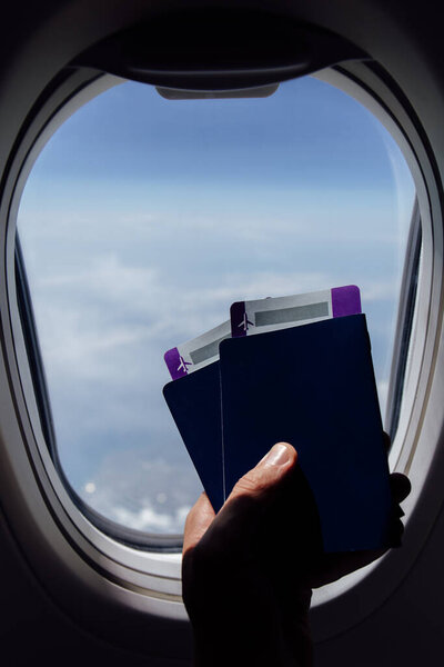 Cropped view of man holding passports with boarding passes near porthole in airplane 