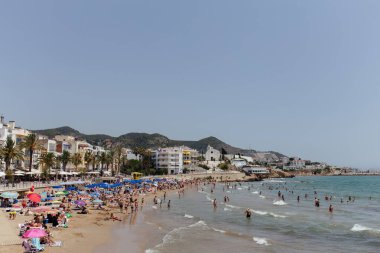 CATALONIA, SPAIN - 30 Nisan 2020: İnsanlar kumlu sahilde dinleniyor ve sahildeki binaların ve palmiye ağaçlarının yakınında denizde yüzüyorlar