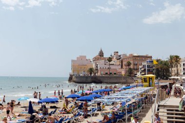 CATALONIA, SPAIN - 30 Nisan 2020: İnsanlar arka planda şemsiyeler, binalar ve mavi gökyüzü ile plajda dinleniyor 