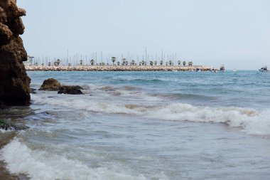 Kıyıda kayalar ve deniz dalgaları, iskele ve arka planda tekneler Katalonya, İspanya 'da 