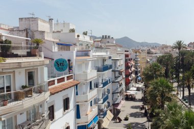CATALONIA, SPAIN - 30 Nisan 2020: Arkaplanda mavi gökyüzü olan binalar, palmiye ağaçları ve dağları olan şehir caddesi 