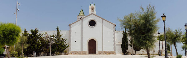 Horizontal crop of trees near white facade of chapel with blue sky at background in Catalonia, Spain 