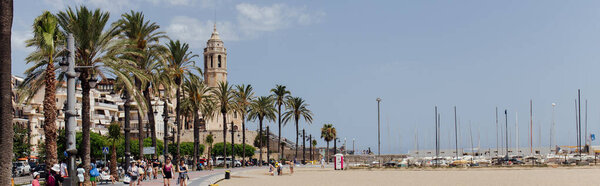 BARCELONA, SPAIN - APRIL 30, 2020: People on urban street with palm trees and Church of Saint Bartolomeus and Santa Tecla at background, panoramic crop 