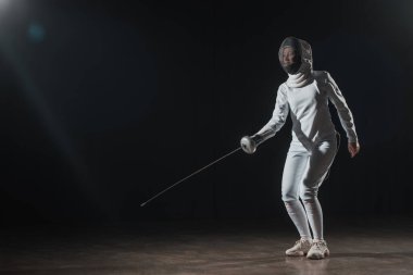 Swordswoman in fencing mask and suit training with rapier under spotlight on black background clipart
