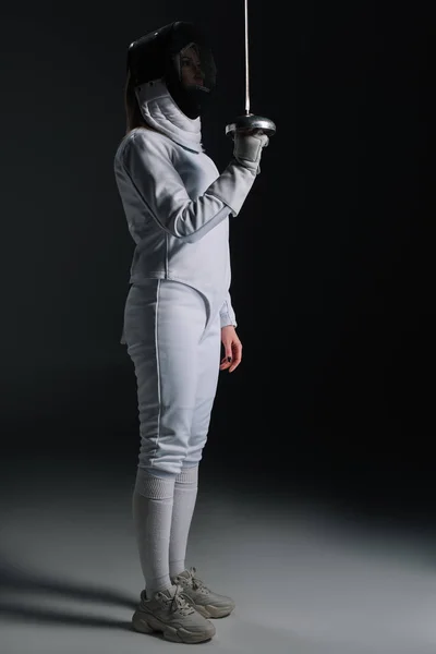 Side view of fencer in fencing suit and mask holding rapier on grey surface on black background