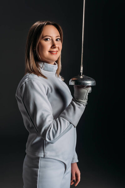 Вид сбоку красивой девушки, улыбающейся перед камерой и держащей в руках рапиру в черном
 