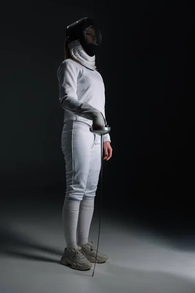 Fencer in fencing suit and mask holding rapier on grey surface on black background