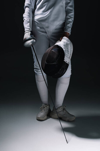 Cropped view of fencer holding rapier and fencing mask on white surface on black background