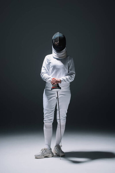 Fencer in fencing mask and suit holding rapier under spotlight on black background