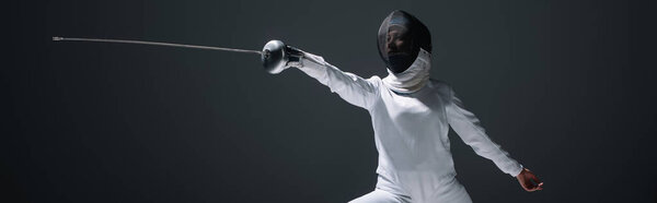 Panoramic shot of fencer in fencing suit and mask exercising isolated on black 