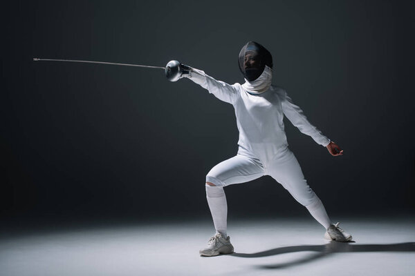 Женщина-фехтовальщица в защитной маске и костюме держит рапиру на белой поверхности на черном фоне
