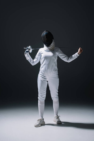 Fencer in fencing mask holding rapier while standing under spotlight on black background