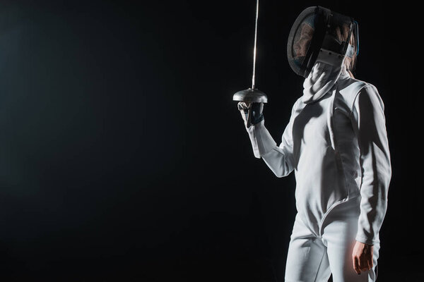 Fencer in fencing mask and suit holding rapier isolated on black 