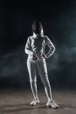 Fencer with hand on hip holding rapier on black background with smoke  clipart