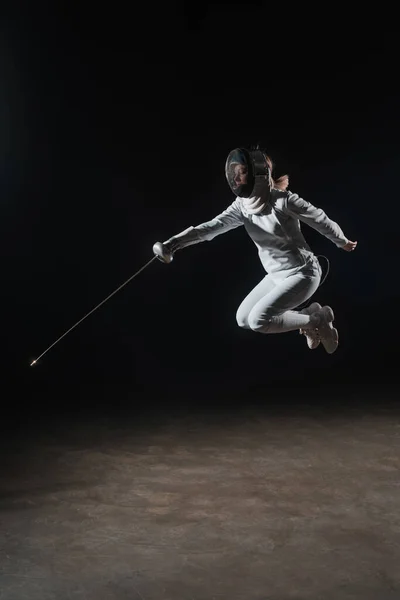 Fencer in fencing mask holding rapier and jumping on black background