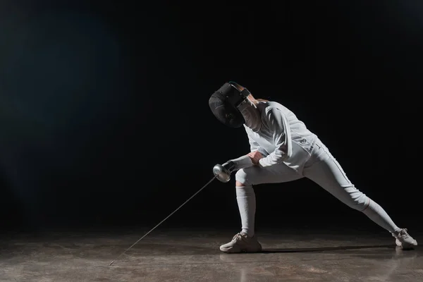 Fencer in fencing mask and suit holding rapier and doing lunge on black background