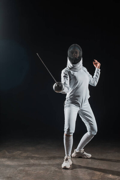 Fencer in fencing mask holding rapier while training under spotlight on black background