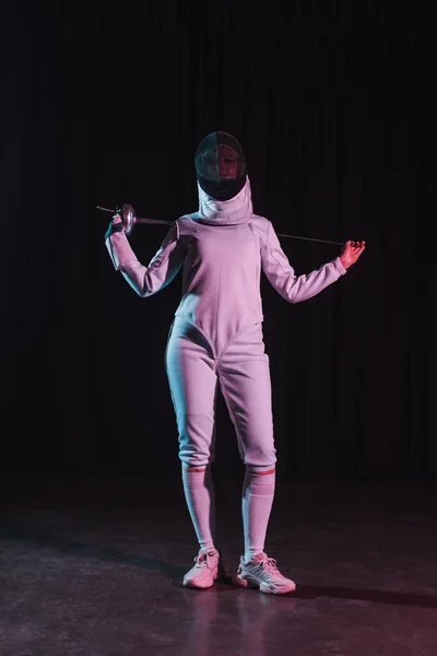 Fencer in fencing mask and suit holding rapier near shoulders on black background