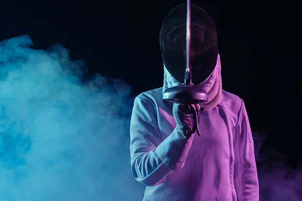 Swordswoman in fencing mask holding rapier on black background with smoke