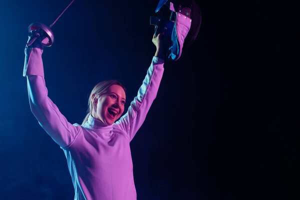 Веселый фехтовальщик, держащий защитную маску и рапира на черном фоне с освещением
 