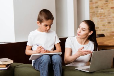Mother point with hand at laptop to son holding color pencil near books on couch  clipart