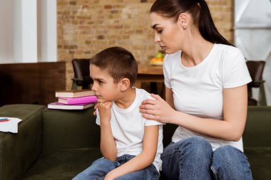 Mother embracing sad son near books on couch  clipart