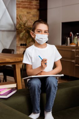 Selective focus of kid in medical mask holding sketchbook and pencil near books on couch  clipart