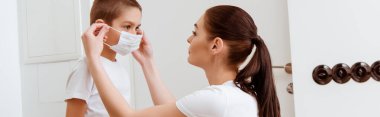 Panoramic shot of mother putting medical mask on son in hallway  clipart