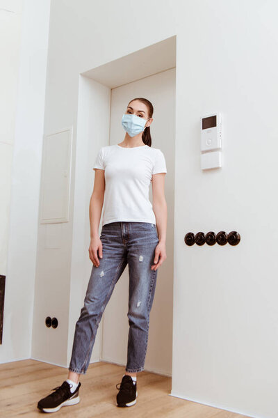 Woman in medical mask looking at camera while standing in hallway at home 