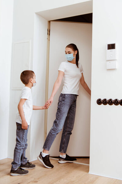 Mother and son in medical masks holding hands near door in hallway