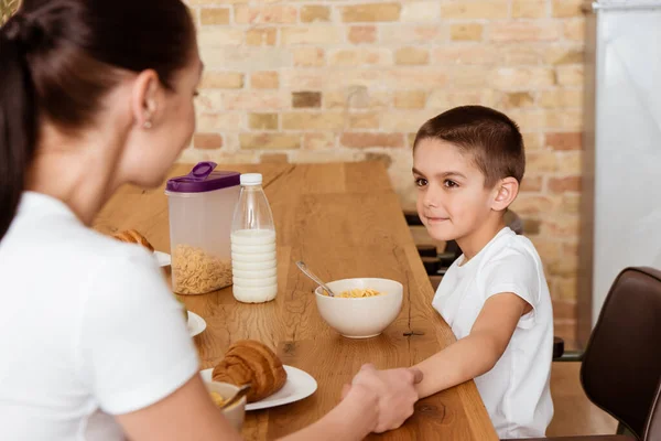 キッチンテーブル上のシリアルやクロワッサンの近くに手を保持男の子と母親の選択的な焦点 — ストック写真