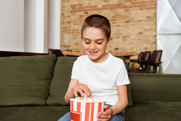Улыбающийся мальчик ест попкорн, сидя на диване в гостиной
