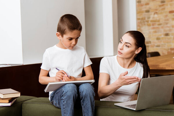 Mother point with hand at laptop to son holding color pencil near books on couch 