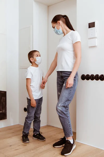 Mother and son in medical masks looking at each other while holding hands in hallway