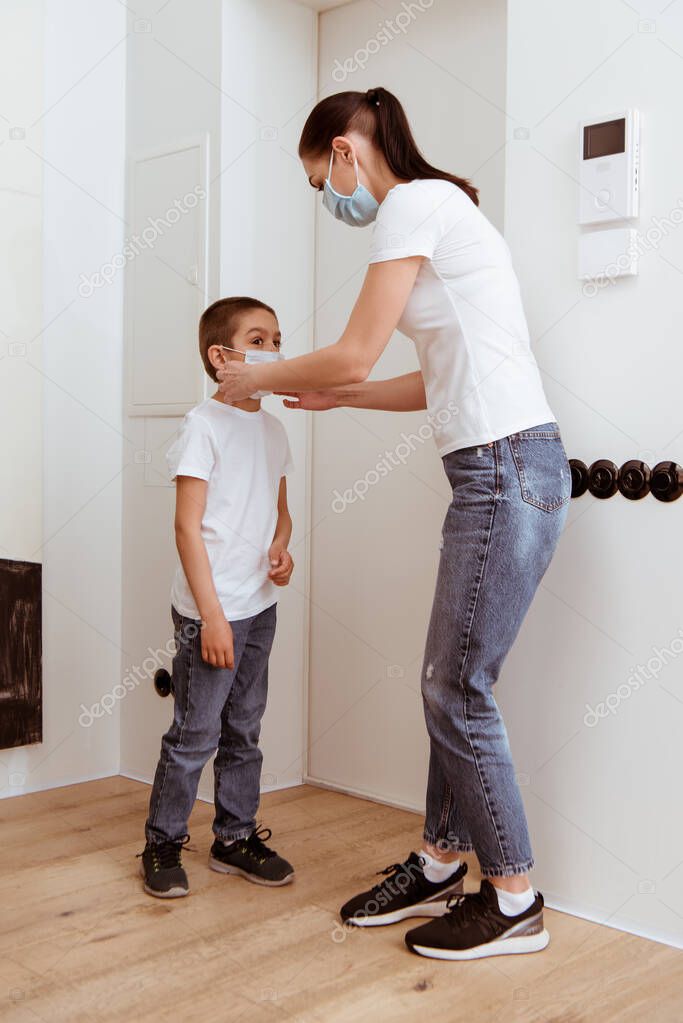 Woman adjusting medical mask on son near door in hallway 