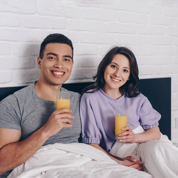 幸せな混合レースマンと陽気な女性がベッドの中でオレンジジュースのグラスを保持 — ストック写真