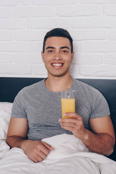 cheerful mixed race man holding glass of orange juice in bed