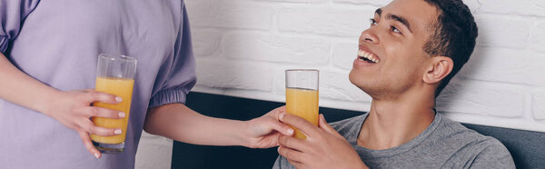panoramic crop of woman giving glass of orange juice to happy mixed race boyfriend in bedroom 