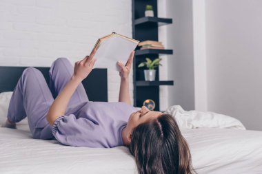 Genç bir kadın yatağa uzanıp kitap okuyor. 