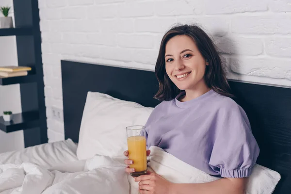 幸せな若い女性はカメラを見てオレンジジュースのグラスをベッドの中で持っています — ストック写真