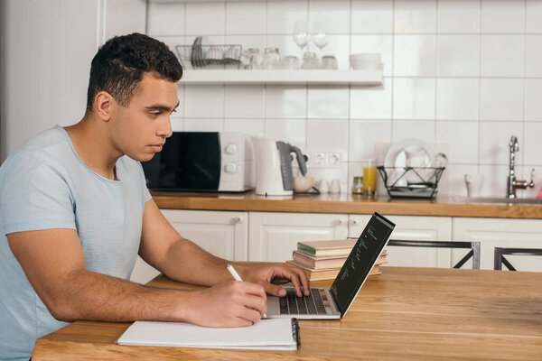 KYIV, UKRAINE - MAY 14, 2020: handsome mixed race man holding pen near notebook and using laptop with javascript 