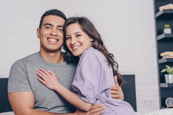 positive mixed race man and cheerful woman hugging at home