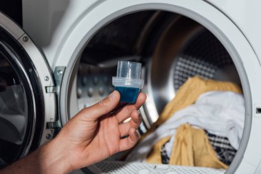 cropped view of man holding measuring cup with detergent near washing machine with dirty clothing  clipart