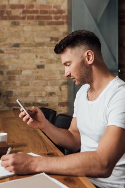 Akıllı telefon kullanan ve mutfaktaki masada deftere yazı yazan adamın yan görüntüsü 