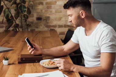 Mutfak masasının yanında erişte yerken akıllı telefon kullanan bir adamın yan görüntüsü. 
