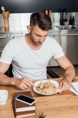 Mutfak masasında akıllı telefonun yanında erişte yiyen bir adamın seçmeli odak noktası. 