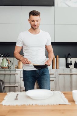 Mutfak masasında çatal ve tabağın yanında tava ve spatula tutan yakışıklı adamın seçici odağı. 