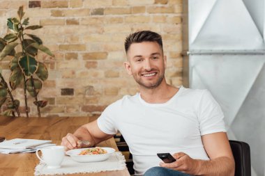 Smiling man holding remote controller near noodles and cup on kitchen table  clipart
