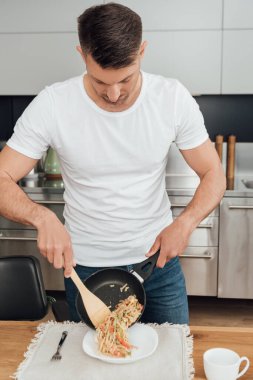 Mutfak masasına erişte dökerken elinde tava ve spatula tutan bir adam. 