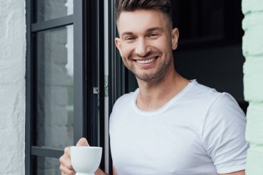 Teras kapısının yanında elinde bir fincan kahve tutarken kameraya gülümseyen yakışıklı adamın seçici odağı 